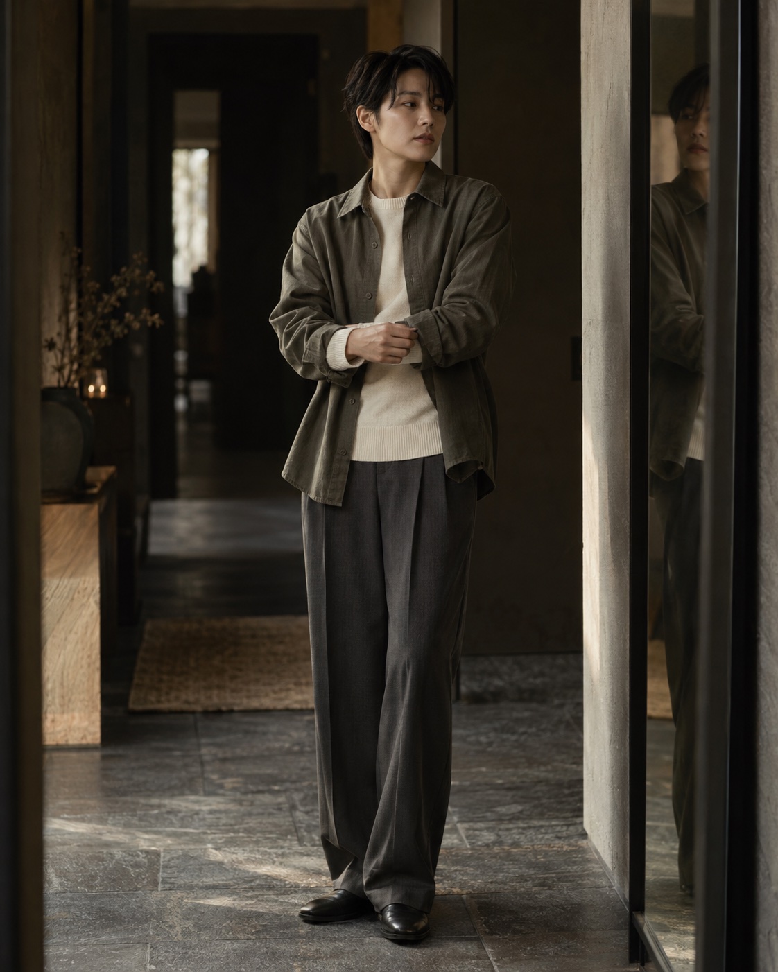 A person adjusting a cuff beside a mirror, wearing a grey-green overshirt and grey trousers